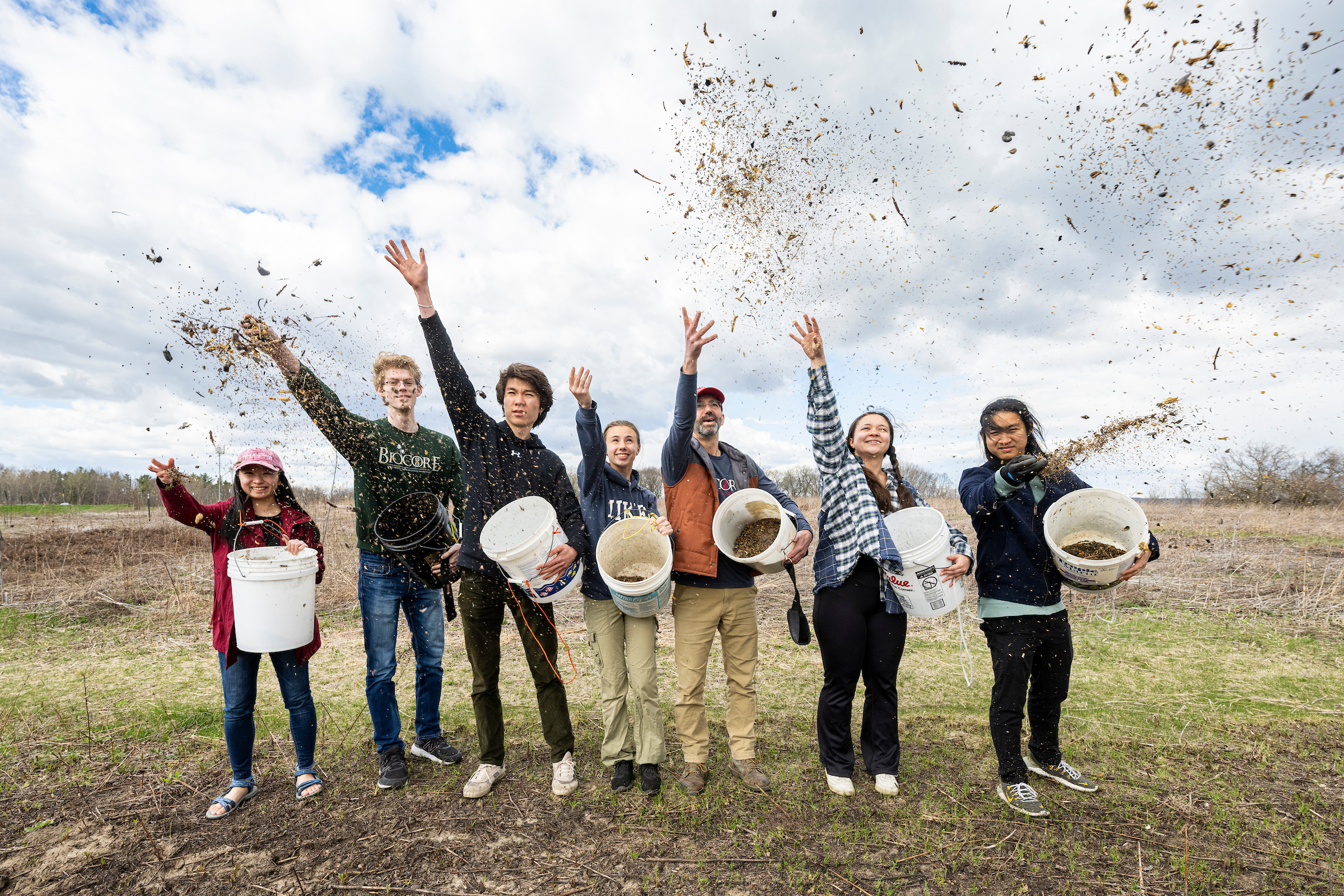 A line of people in a field have one arm thrown up into the sky, releasing the handful of seeds they just gathered from the large white buckets held in their other arm.